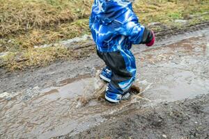 un niño camina abajo el Arroyo en el primavera después el nieve se derrite en el primavera. un chico en un azul mono y caucho botas en un charco. foto