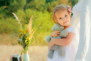 Little beautiful girl in a white dress gray bear hugs. The girl on the background of the tent tilted head. A child's birthday is on the street. photo