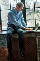 A man looks at the laptop screen and writes the message. The man is sitting on a windowsill near the window. Freelancer works at home in a comfortable environment. Soft focus photo