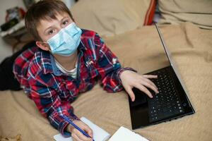 boy in protective medical mask lies near laptop and writes in work book. child does lessons watching online broadcast lessons on Internet. e-learning, remote studying internet concept photo