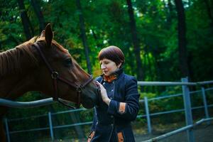 A woman in a blue long coat and with short dark brown hair extended her hand to stroke the horse. The horse pricked up his ears. A train for horses against the background of a green forest. photo