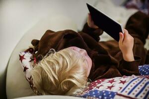 A little girl lies on a soft couch in a fluffy home jumpsuit with a tablet in her hands. The child focuses and enthusiastically looks at the tablet screen and plays games. photo