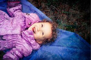 un pequeño niña mentiras en el suelo y carcajadas con alegría. un niña con corto Rizado pelo sonrisas felizmente. un contento niño camina en un cobija para un caminar. foto