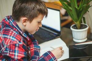 boy writes homework in notebook sitting near open laptop and lying tablet on table photo