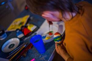 un niño decora un Pascua de Resurrección huevo en el colores de el arcoíris. un niño sostiene un huevo y pinturas eso con un cepillar. preparando para el celebracion de Pascua de Resurrección. foto