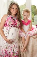 Pregnant woman with little beautiful daughter sitting on the windowsill. Woman and girl in pink dresses. photo