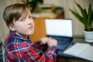 boy teaches lessons sitting at home by laptop. working notebook on student's desk during distance learning on coronavirus quarantine. child turned around photo