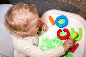 baby holds mold for playing with sand at home. Educational toys for children up to year. girl plays with kinetic sand. child builds shepherds in form of letters of sand. Early development of children. photo