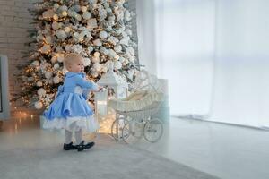 Baby girl wearing cute dress and headband, carries stroller in festively decorated room with garland of lights. photo