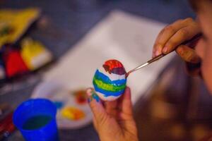 un niño decora un Pascua de Resurrección huevo en el colores de el arcoíris. un niño sostiene un huevo y pinturas eso con un cepillar. preparando para el celebracion de Pascua de Resurrección. foto