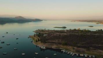 aéreo zangão vôo sobre uma lago, água barragem, com montanhas, e barcos em litoral, às nascer do sol. lindo sonhadores panorama visualizar. video