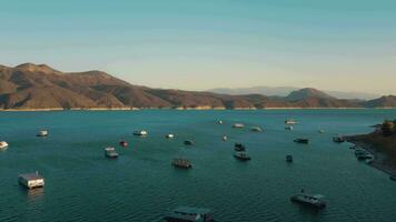 aéreo zumbido volador terminado un lago, agua presa, con montañas, y barcos en línea costera, a amanecer. hermoso soñador paisaje vista. video