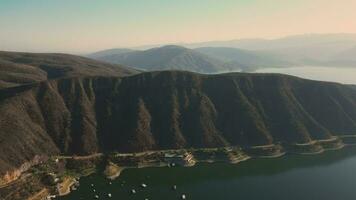 aéreo zumbido volador terminado un lago, agua presa, con montañas, y barcos en línea costera, a amanecer. hermoso soñador paisaje vista. video