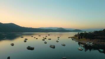 aéreo zangão vôo sobre uma lago, água barragem, com montanhas, e barcos em litoral, às nascer do sol. lindo sonhadores panorama visualizar. video