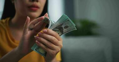 cenas fechar acima mãos do jovem ásia mulher apreciar contando dinheiro dólares notas em sofá dentro a vivo quarto às lar. video