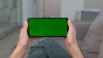 een vrouw houdt een telefoon met een groen scherm in een horizontaal positie met beide handen terwijl aan het liegen Aan de sofa Bij huis. video