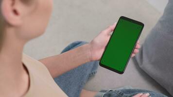 mulher tocando em a verde tela do dela telefone enquanto sentado em a sofá às lar. tocando em Centro tela video