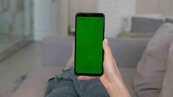 A woman makes a swipe down gesture on the green screen of a smartphone while lying on the sofa at home. video