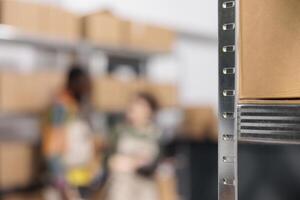 Selective focus of metallic shelves full with cardboard boxes, in background diverse workers discussing goods inventory report. Storage room team working at customers orders, preparing packages photo