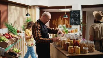 Portrait of elderly vendor working in zero waste organic food supermarket, adding pantry staples on shelves. Older employee restocks local grocery store with bulk products in biodegradable jars video