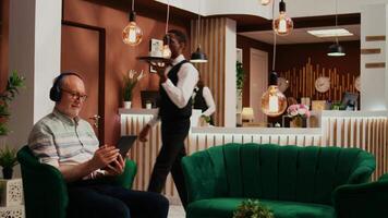 hotel invitado teniendo divertido con tableta en vestíbulo, acecho película o televisión espectáculo antes de registrando a frente escritorio recepción. mayor persona disfrutando película en transmisión servicios en línea, utilizando dispositivo. video