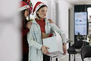 Asian employee upset by company manager leaving Christmas decorated office after deciding to quit. Team leader holding desk belongings box, bidding farewell to assistant before starting another job photo