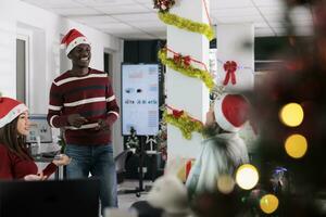 Multiethnic business team enjoying job shift break, bonding together in festive decorated office during winter holiday season. Coworkers feeling Christmas spirit mood, relaxing in xmas adorn workspace photo