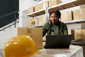 African american employee checking warehouse logistics on laptop computer, preparing clients orders for shipping. Storage room supervisor working at merchandise inventory in storehouse photo