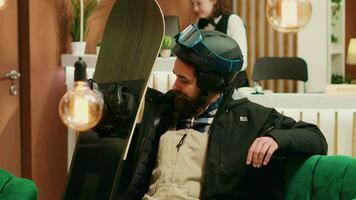 Athletic traveler seated in lobby looking at his snowboard and feeling excited to start winter sport on mountain slopes. Young man equipped with helmet and goggles ready for wintertime leisure. video