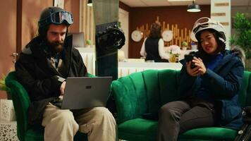 Focused couple using gadgets in lobby, arriving at ski lodging during wintertime season. Man searches for slope activity online while woman checks social media messages. Handheld shot. video