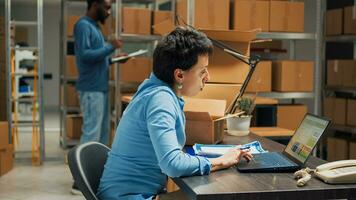 Entrepreneur using laptop to plan storage logistics, preparing merchandise order to ship to customers. Young employee putting products in packages, taking notes for stock inventory. photo