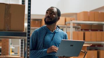 African american man working on merchandise inventory using laptop and taking notes in storehouse. Entrepreneur checking products in cardboard boxes, working on quality control and distribution. photo