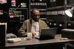 Private investigator sitting at desk and working on laptop in agency at night. Concentrated african american criminalist searching crime case file on computer in police detective office photo