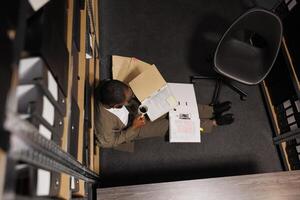 Criminologist drinking coffee and reading crime case on floor. Police investigator holding mug and folder with report papers while studying archival records at night time top view photo