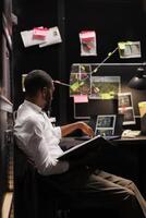 Policeman stuying crime case and analyzing photos on laptop, sitting at desk with evidence scheme board on wall. Investigator checking surveillance camera photographs on computer