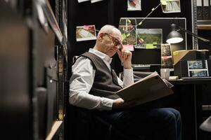 Old police officer working overtime at missing person case, analyzing crime scene evidence. Elderly private detective sitting at desk reading confidential victim files in arhive room photo