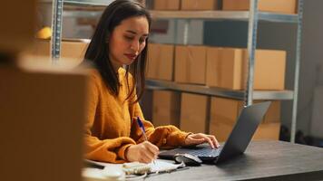 Female entrepreneur taking notes on papers and pc, working in warehouse to do inventory and shipping logistics. Young adult checking products in boxes before distribution. Handheld shot. photo