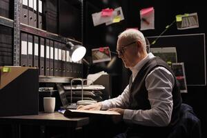 Elderly police officer analyzing confidential federal files, working at criminal investigations in arhive room. Senior private detective checking missing person case, looking at victim report photo