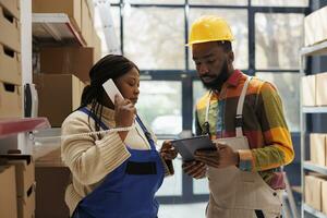 Warehouse manager calling in logistics office and asking about inventory report. African american storehouse workers team looking at stock information on tablet and talking on telephone photo