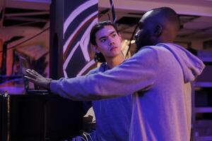 Hackers discussing malware code on laptop while planning illegal activities. African american and asian criminals talking about online server hacking technique in abandoned warehouse photo
