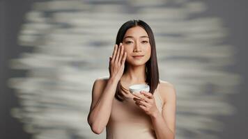 Glowing asian lady applying moisturizing face cream to promote skincare routine and uplifting products. Flawless woman using facial serum and moisturizer for new dermatology ad campaign. photo