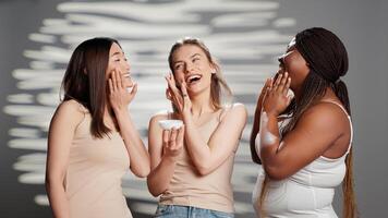 grupo de perfecto señoras aplicando hidratante cara crema en estudio, promoviendo luminoso protección de la piel productos interracial joven mujer utilizando hidratante y suero para nuevo dermatología campaña. foto