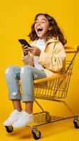 Excited happy girl using smartphone in shopping cart trolley on yellow background AI Generated photo