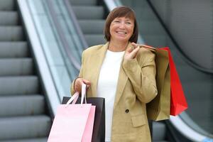 elegante sonriente adulto mujer es levantamiento abajo en escalera mecánica con vistoso compras bolsas. atractivo hembra gasto ocio hora a grande moderno centro comercial. en línea compras concepto, estacional ventas.negro viernes foto