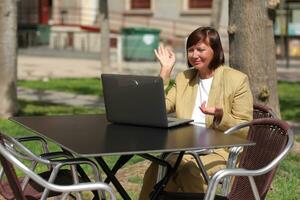 Stylish adult businesswoman, freelancer, mentor talks by smartphone and browsing laptop at city street cafe. Concept of remotely working, leadership, empowerment of mature women in business photo