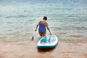 Back view of adult overweight woman in swimsuit with SUP board and paddle goes into sea. Wellness middle aged female enjoys surfing on paddle board on summer vacation. Active modern lifestyle. photo