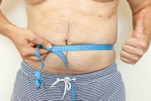 Man measures his fat belly with measuring tape and shows thumb up. Concept of weight loss, health problems of obese people. Controlling eating and active lifestyle. World Obesity Day photo