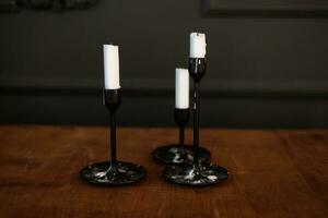 Modern three black glass candlesticks with white candles on wooden table in antique style on dark wall background. Minimalist elegant decor with candle holders with wax different size. photo