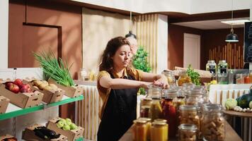 Portrait of smiling woman working in zero waste supermarket, adding pantry staples on shelves. Cheerful merchant restocks local grocery store with organic food in reusable jars, close up video