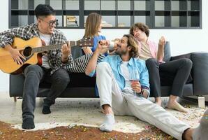 grupo de joven multirracial personas teniendo divertido a casa fiesta. joven amigos jugando guitarra y canto mientras sentado en sofá en vivo habitación. foto